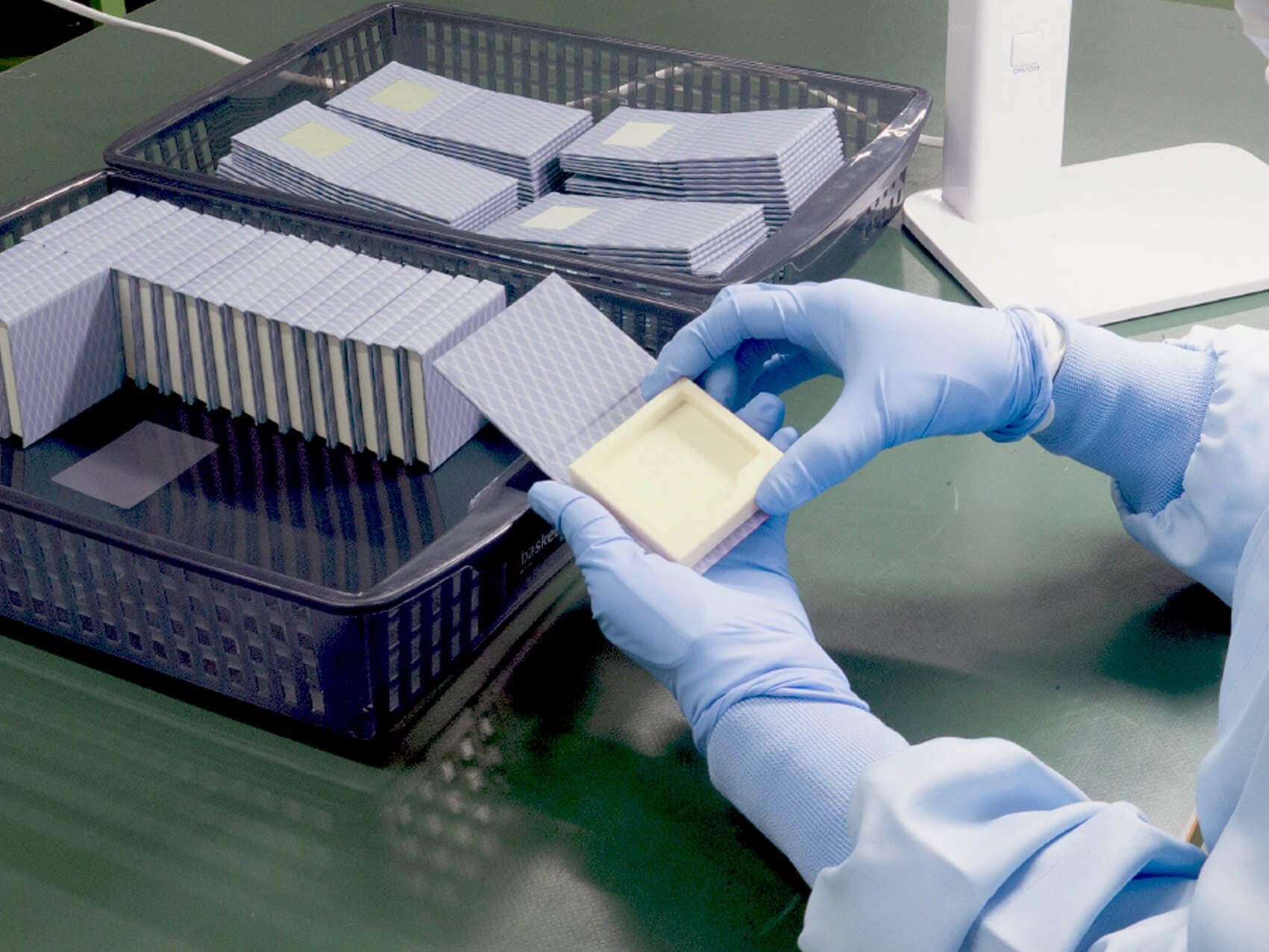 Gloved hands holding a pale yellow square mold near stacked blue grid-patterned samples in a lab tray.